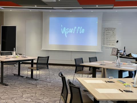 Salle de séminaire vide avant l'arrivée du groupe : tables en îlots, verres retournés, écran projeté et agenda manuscrit sur paperboard.